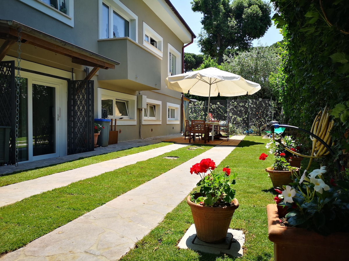 A private garden features well-maintained lawn and flower pots with vibrant blooms. Outdoor furniture, including a large umbrella, is arranged around a table, providing a comfortable space for dining or relaxation. The garden is bordered by greenery, creating a serene environment.