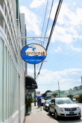 The image shows a sign for 'Shorebreak' hanging above the sidewalk. The background features a partly cloudy sky, with pedestrians and parked cars visible along the street, providing a glimpse of the vibrant beach community atmosphere.
