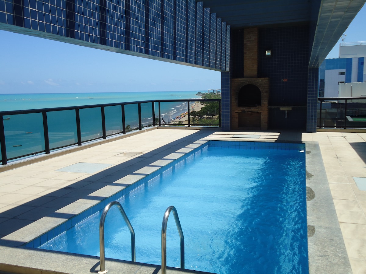 A private outdoor pool is positioned on a spacious terrace, offering views of the ocean. The pool is surrounded by tiled flooring and features a sliding ladder for entry. A barbecue area is located nearby, complemented by blue tiles on the upper section of the terrace.