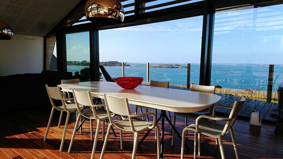 A spacious dining area is presented with a long table surrounded by eight light-colored chairs. A red bowl is positioned at the center of the table. Floor-to-ceiling windows offer a panoramic view of the sea, allowing natural light to flood the space.