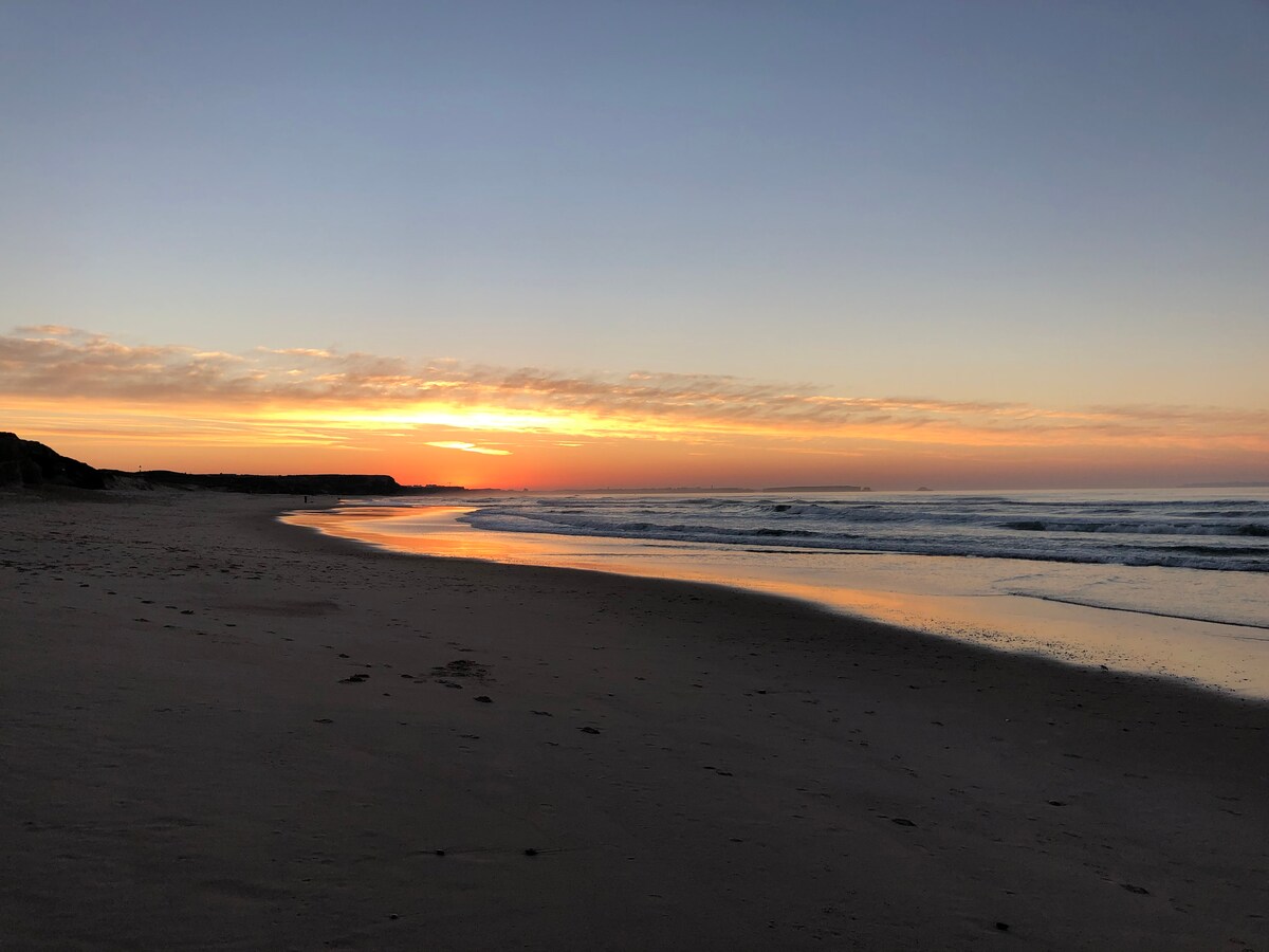 A tranquil beach scene features gentle waves lapping at the shore under a colorful sunset. Soft sands stretch along the curving coastline, while the sky displays hues of orange and pink. Silhouettes of distant cliffs can be seen against the fading light.