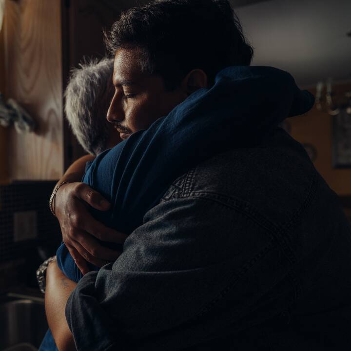 A man with dark hair and a mustache warmly hugs his mother, who has short gray hair. They are in a dim kitchen.