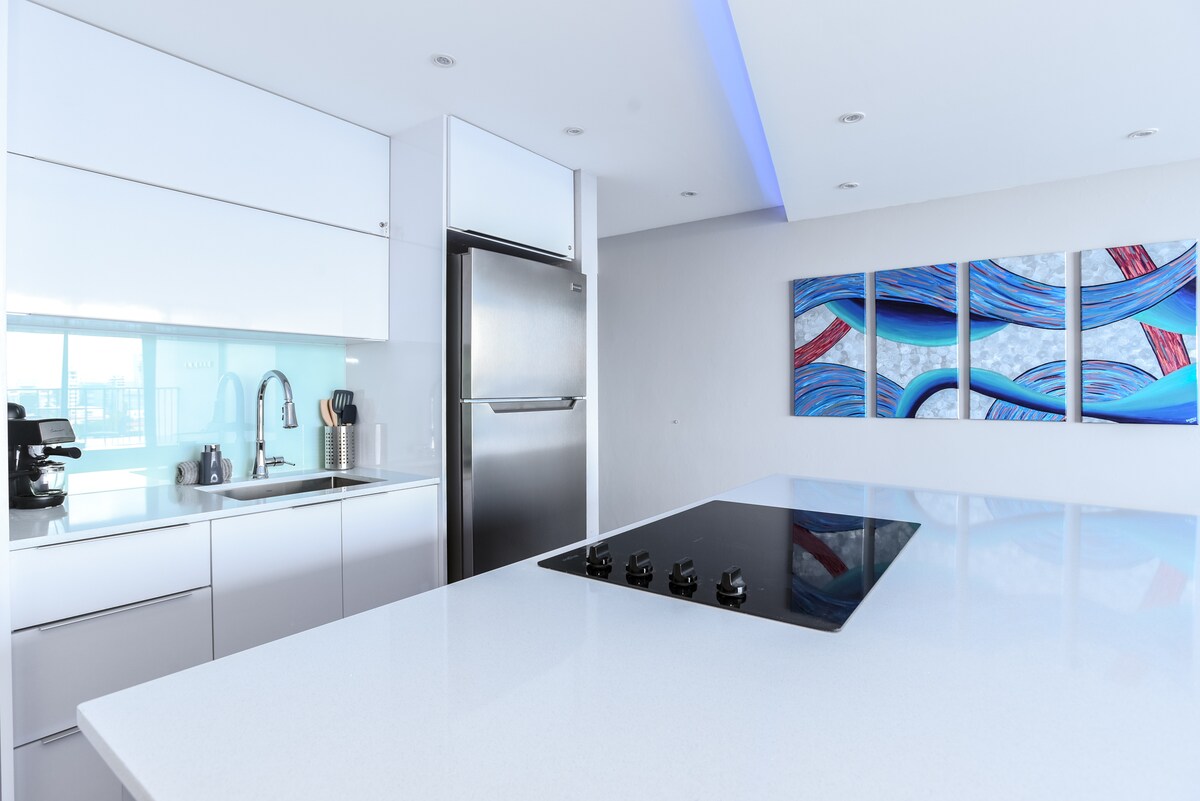 A modern kitchen boasts sleek white cabinetry and a spacious island with a built-in stovetop. A stainless steel refrigerator sits adjacent to the sink area, and contemporary artwork enhances the light-filled space.