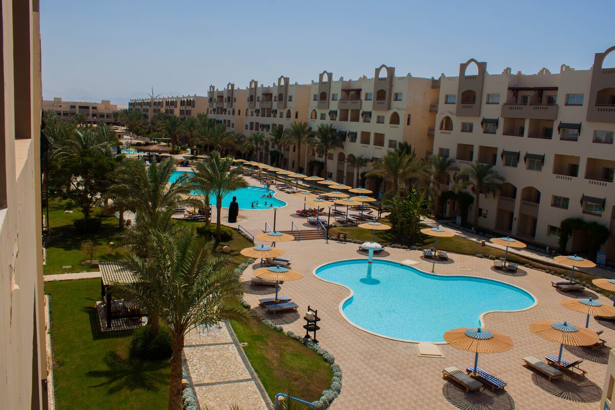 Aerial view of a resort featuring several swimming pools surrounded by lush greenery and palm trees. Sun loungers are arranged around the pools, complemented by sun umbrellas. The architectural layout of the buildings provides a structured yet inviting atmosphere.