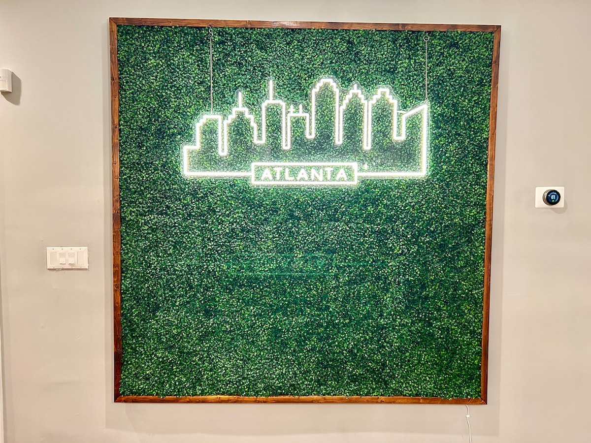 A vibrant faux boxwood wall features a neon outline of the Atlanta skyline, with the city name 'ATLANTA' illuminated in light. The greenery adds a refreshing touch to the wall, creating an engaging focal point in the space.