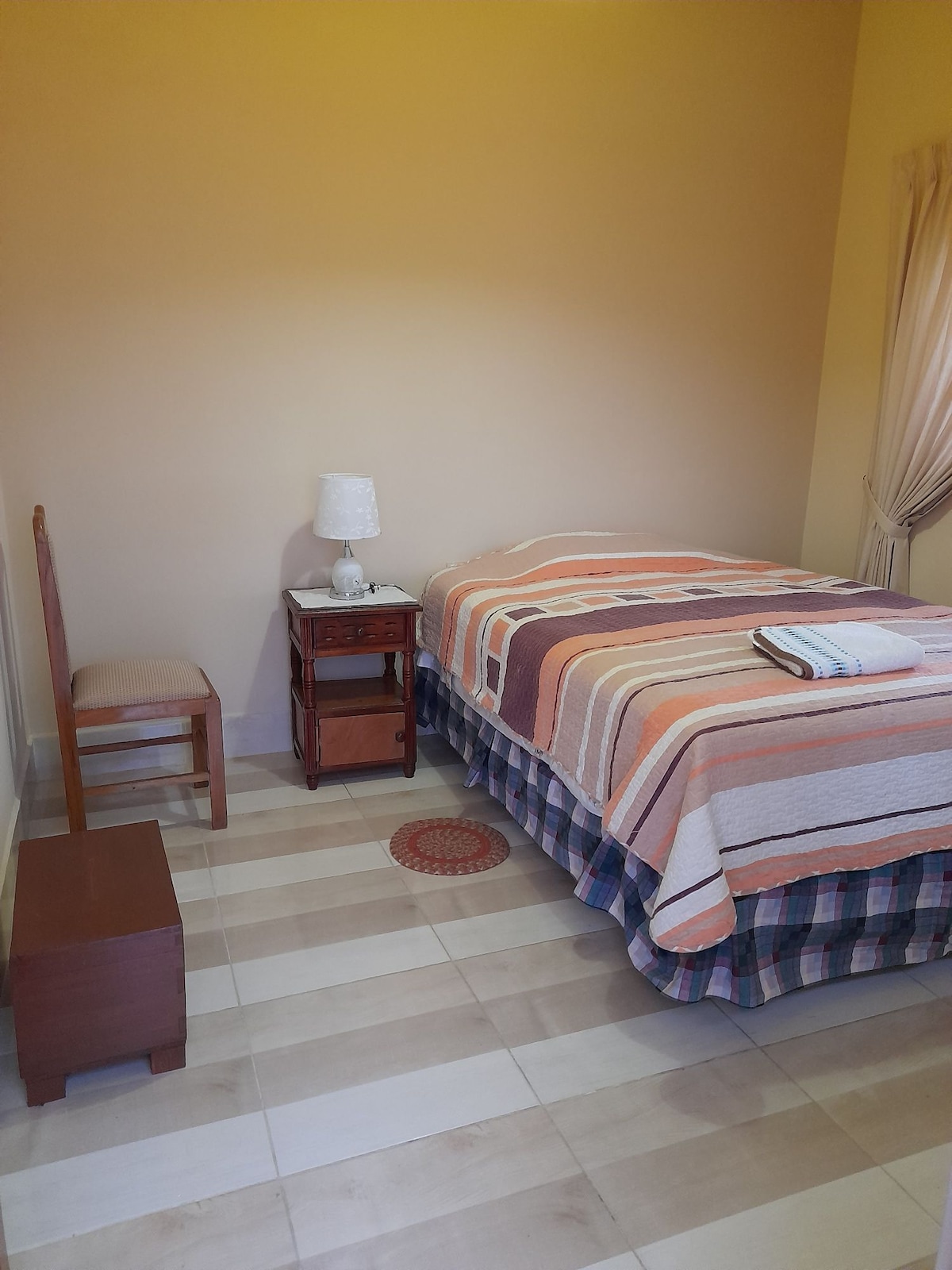 A cozy sleeping area features a striped bedspread atop a spacious bed. A bedside table holds a small lamp, while a chair is placed nearby. Soft light enters through a window draped with sheer curtains, illuminating the warm-toned walls and tiled floor.