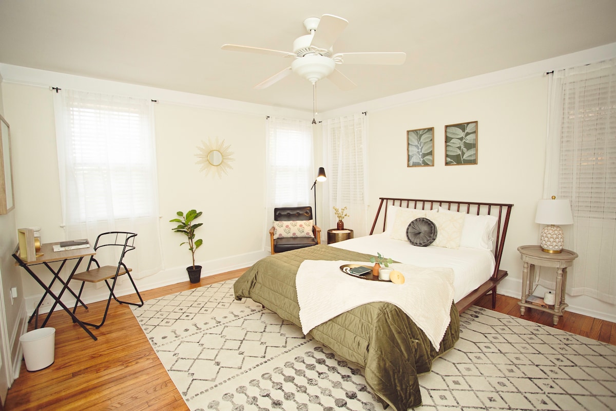 A bright bedroom features a spacious bed adorned with a textured green blanket. Natural light filters through sheer curtains, illuminating an accent chair and a small desk with a chair. Decorative plant elements and framed art pieces add subtle touches to the inviting space.