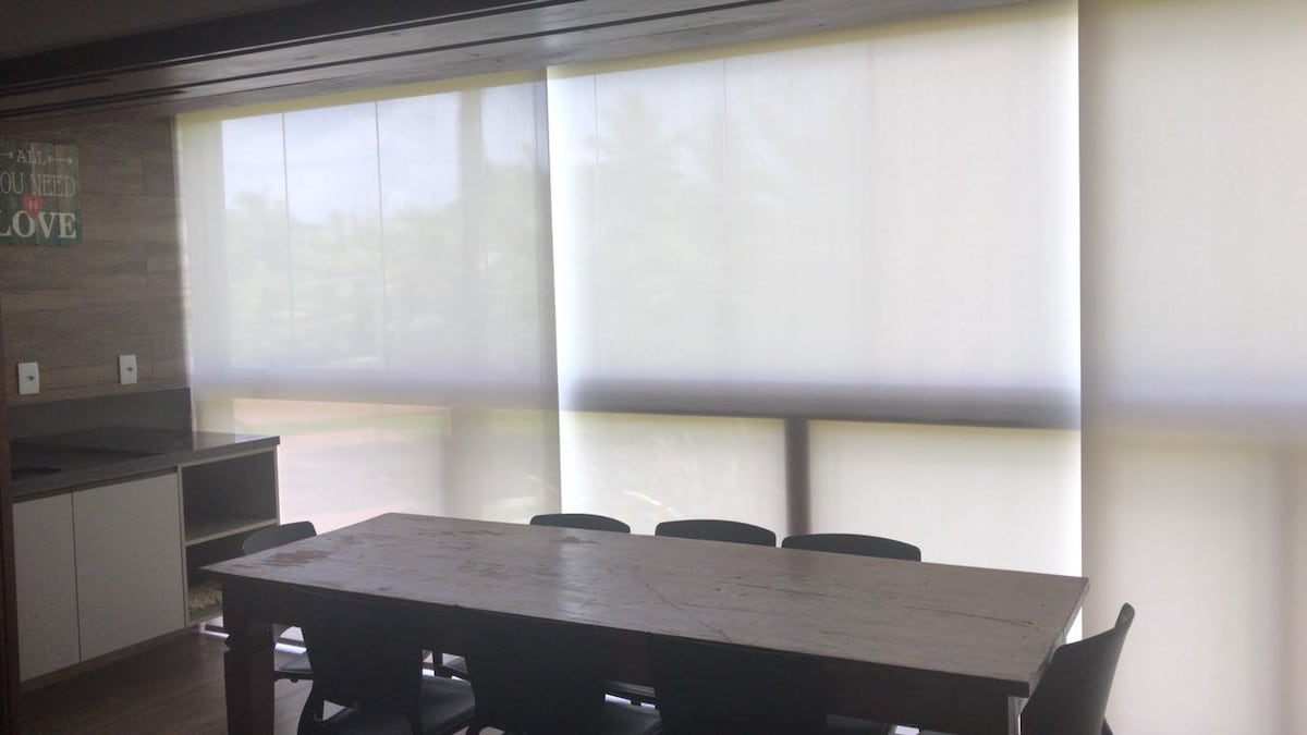 A spacious dining area features a large wooden table surrounded by black chairs. Natural light filters through sheer white window coverings, creating a soft ambiance. Minimalist décor is present on the walls, contributing to a relaxed setting.