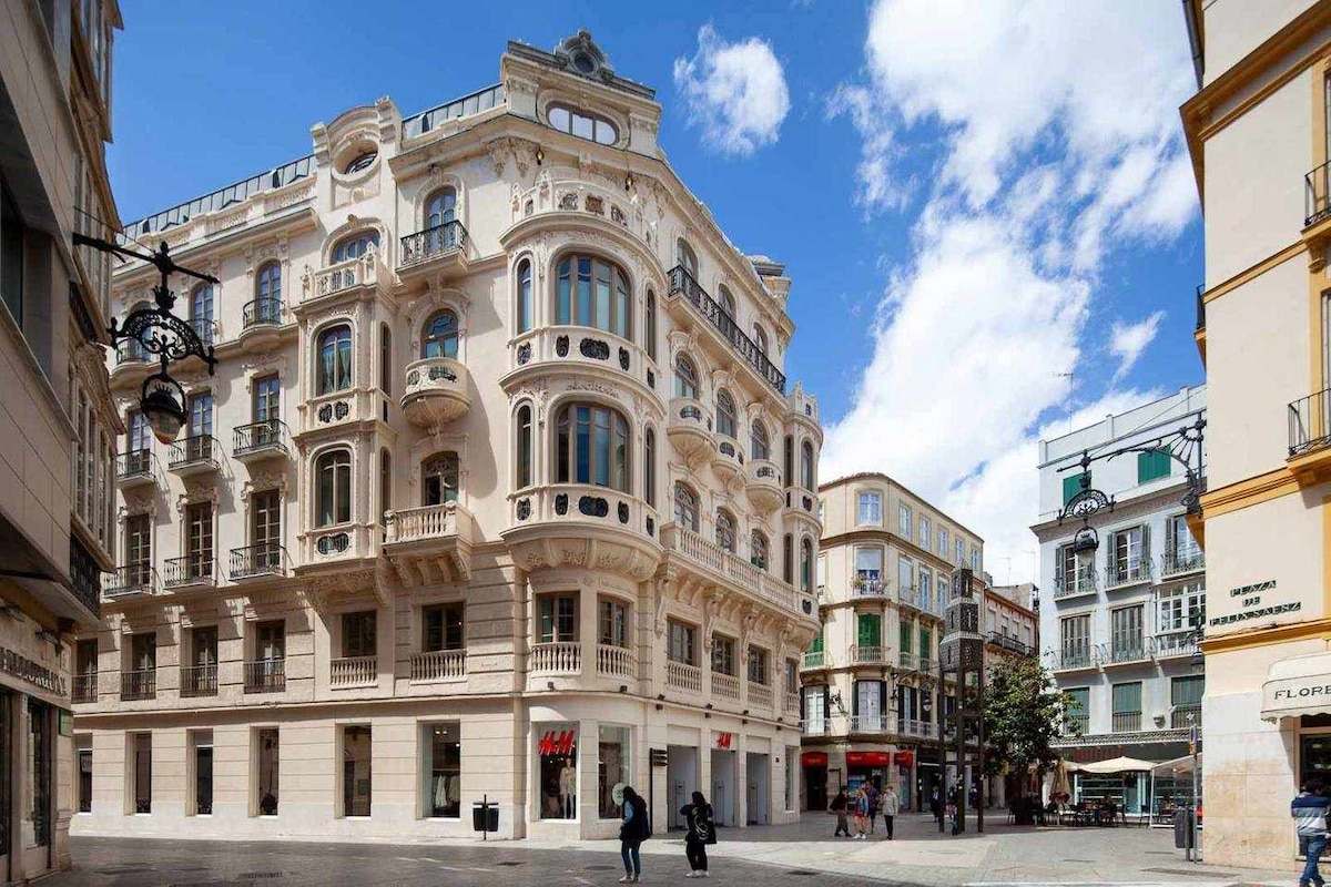 The exterior of a stylish building is showcased, featuring elegant architectural details and large windows. The structure is located at a lively corner, with shops and pedestrians visible on the street. The sky is bright with clouds, adding to the vibrant city atmosphere.
