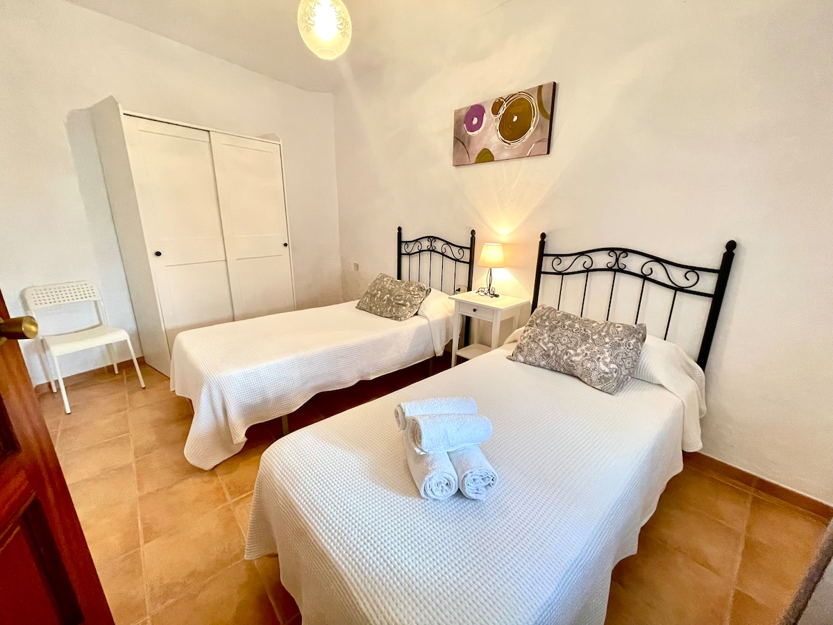 The room features two single beds with light bedding, complemented by decorative pillows. A small bedside table is positioned between the beds, topped with a lamp. A white wardrobe provides storage, while a chair is visible in the corner against a neutral wall.
