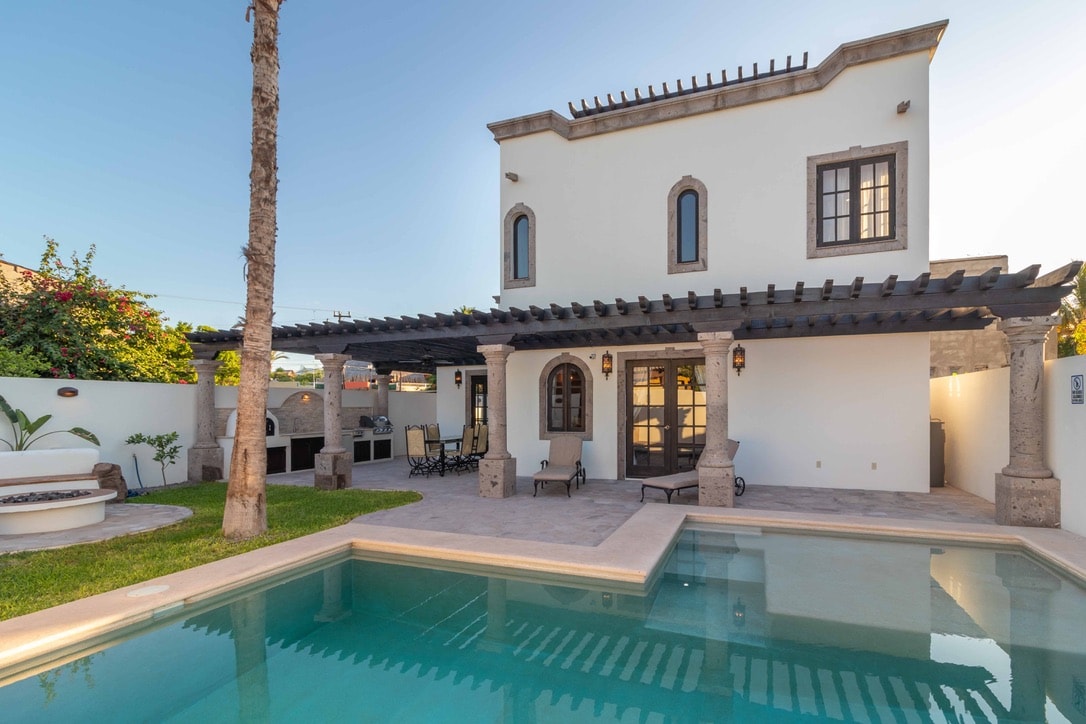 A two-story building is shown with a classic design, featuring multiple windows and a shaded patio area. A sparkling pool is visible in the foreground, surrounded by manicured grass. The outdoor seating area offers a relaxing space under a pergola with decorative lighting.