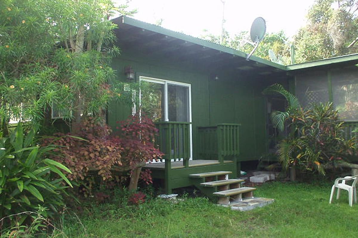 A quaint green cottage is surrounded by lush vegetation and tropical plants. A small set of wooden steps leads up to a front deck, while a satellite dish is mounted on the roof. The entrance features a large window, allowing natural light to enter.