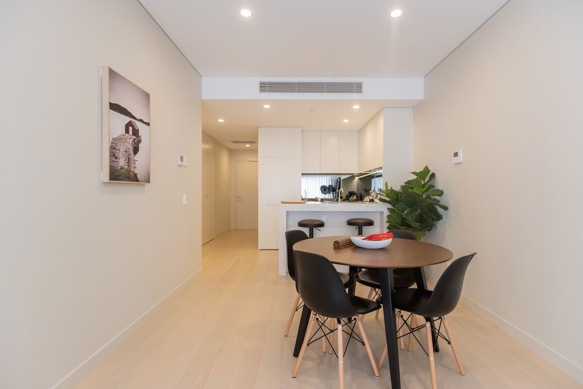A modern dining area features a round table surrounded by four black chairs with wooden legs. A large plant is positioned in the corner, complementing the light-colored wooden flooring. A wall-mounted artwork adds a touch of style, while the open layout leads to a sleek kitchen.