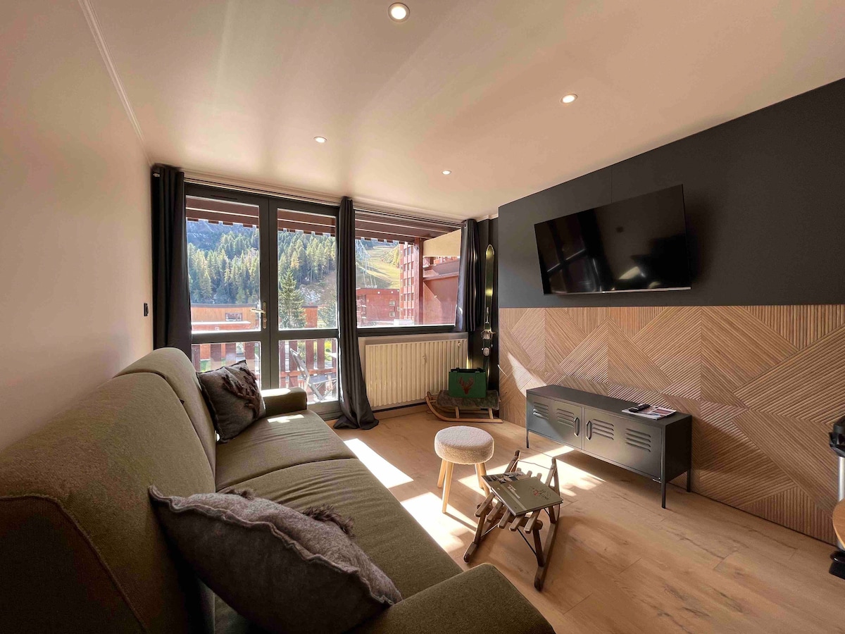 A cozy living area is highlighted by large windows that invite natural light and showcase views of surrounding mountains. A spacious green sofa faces a modern TV mounted on a textured wood accent wall, complemented by a small table and a decorative element.