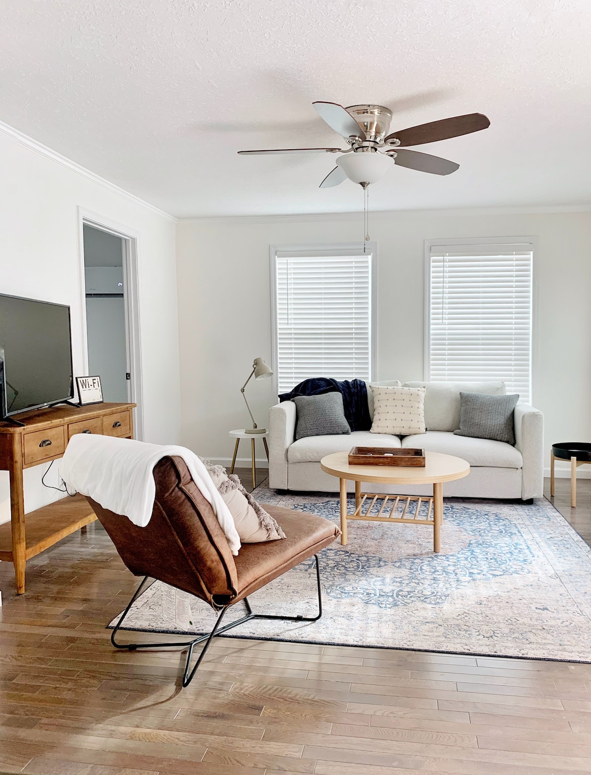 A light-filled living space features a comfortable sofa, a round coffee table, and a stylish armchair. Two windows let in natural light, and a subtle area rug adds warmth to the hardwood floor. A flat-screen TV is positioned against the wall, complemented by a small side table.