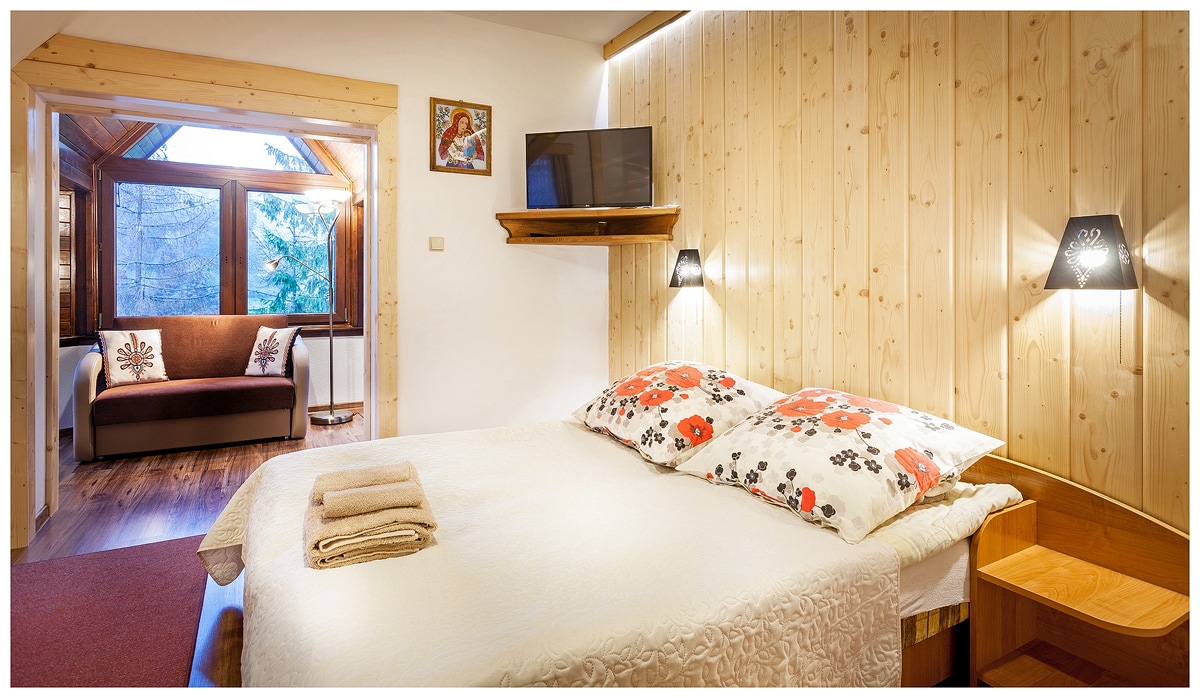 A cozy sleeping area features a spacious bed with floral pillows and a warm quilt. Soft lighting is provided by wall sconces, highlighting the wooden paneling. A separate seating area with large windows offers views of the outdoors, enhancing the room's inviting ambiance.