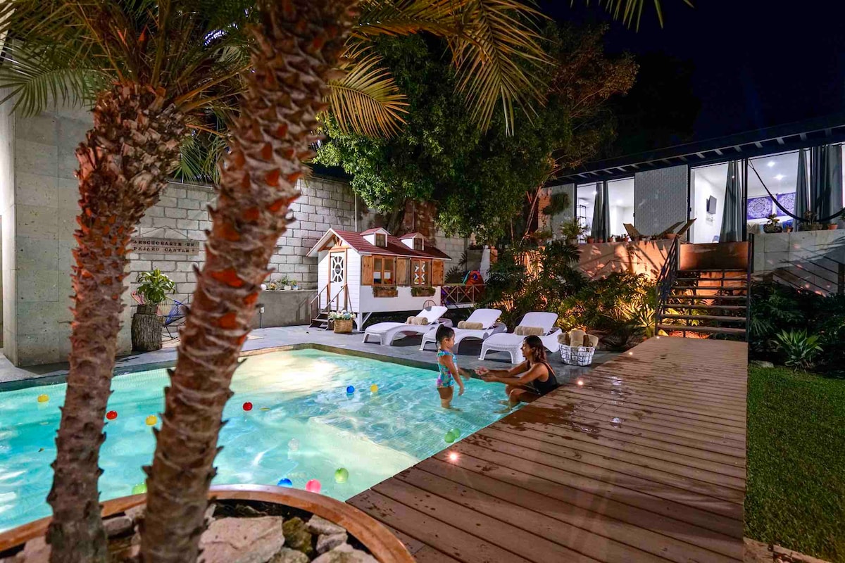 A well-lit backyard features a heated pool surrounded by lounge chairs. A treehouse stands nearby, with playful children enjoying the water under the night sky. Lush vegetation enhances the tranquil outdoor setting, complemented by gentle lighting from the house.