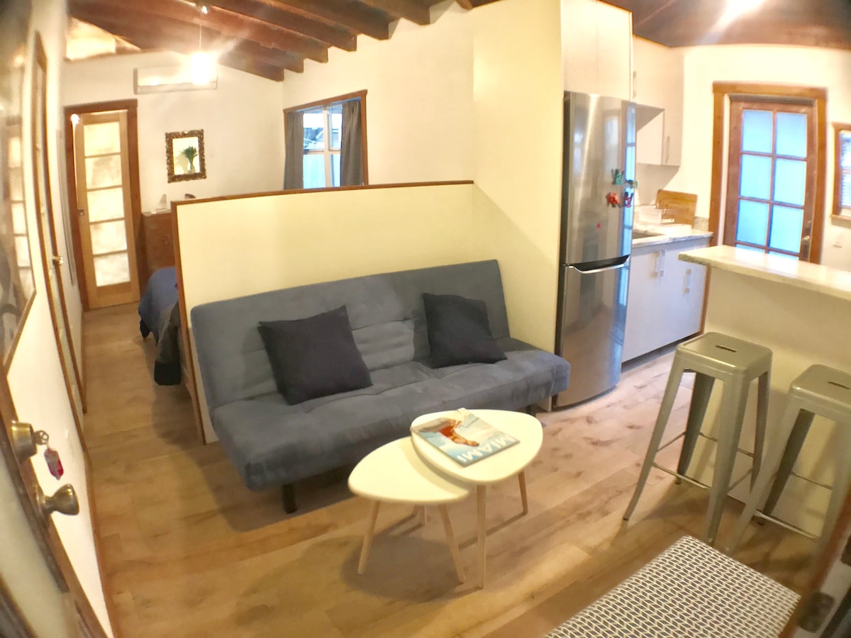 An open living space showcases a blue futon adorned with two black cushions. A small coffee table sits beside it, and modern bar stools are positioned near the kitchen counter. A stainless steel refrigerator and light-colored cabinetry are visible, along with a soft ambiance created by the natural light through the windows.