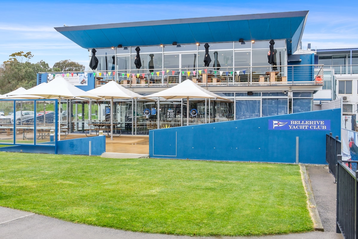 A modern structure of the Bellerive Yacht Club is presented with a blue exterior and large windows. Outdoor seating is arranged under umbrellas with a grassy area in front. Colorful flags are displayed, adding a festive touch to the setting.