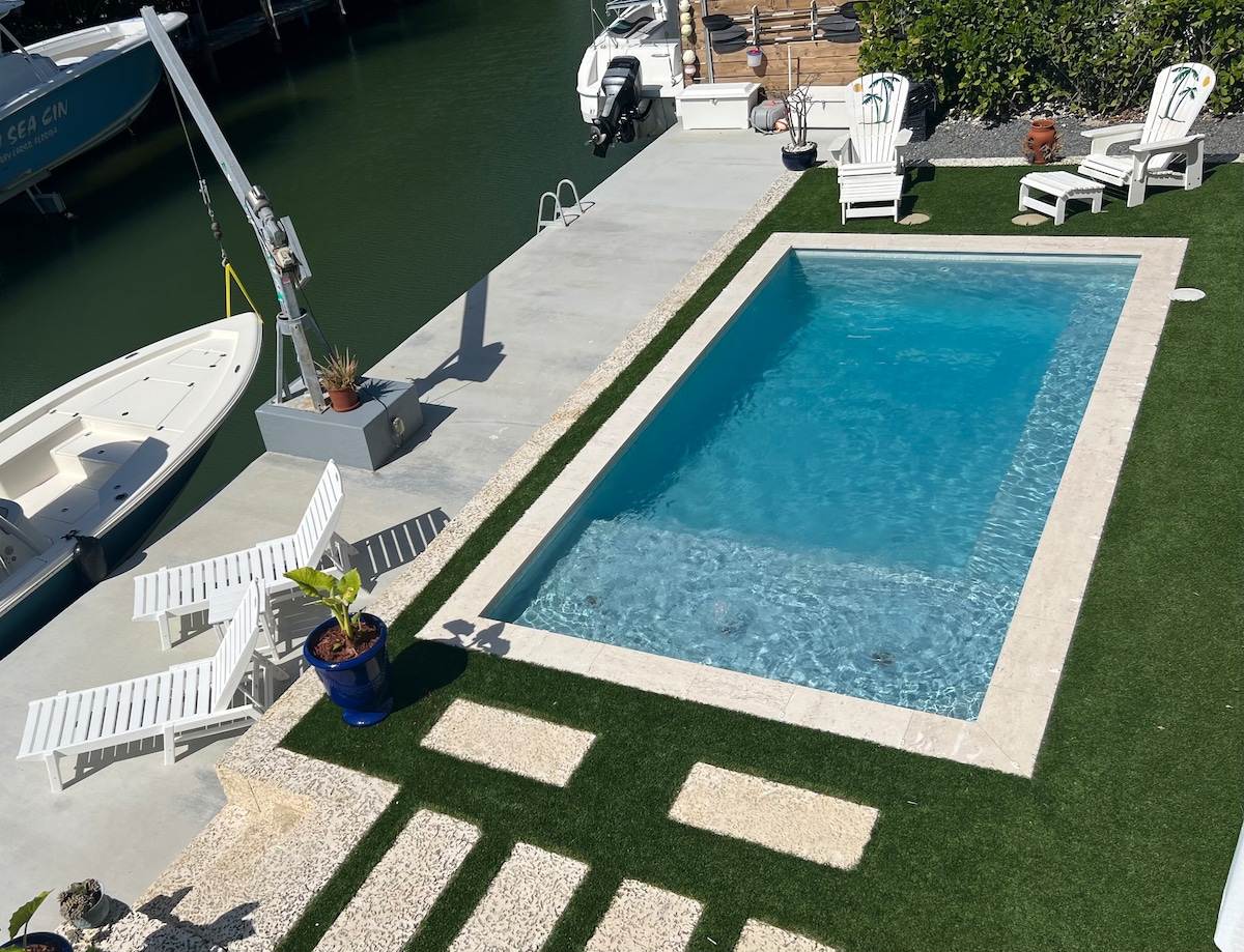 A private pool is surrounded by lounge chairs and greenery. A concrete dock is visible nearby, providing access to the canal. The water in the pool is clear, reflecting sunlight, creating a serene setting for relaxation.