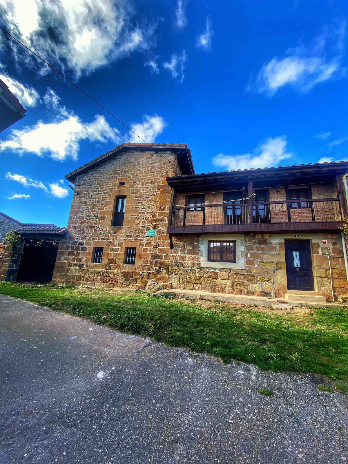 A rustic stone house with two levels is set against a vibrant blue sky. It features a wooden balcony on the upper floor, while large windows allow natural light to permeate the interior. A grassy area surrounds the base, complementing the natural setting.