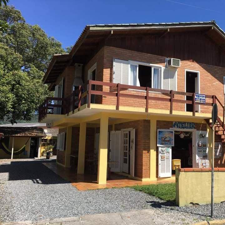 A two-story brick house is shown, featuring a spacious balcony with wooden railings. The entrance is framed by large windows, and the ground floor includes an area with outdoor seating. Lush green trees are visible in the background, providing a natural setting.
