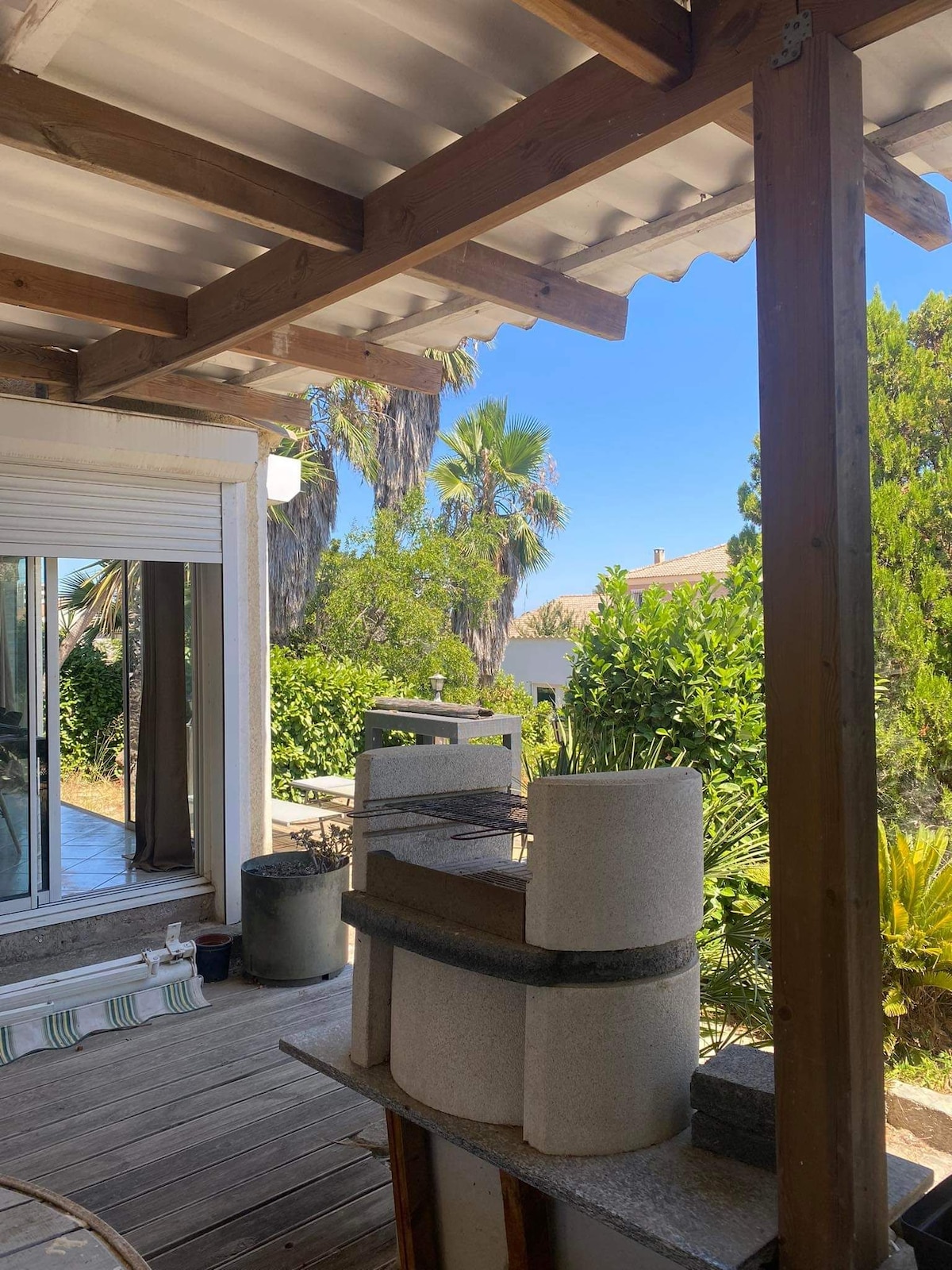 An outdoor kitchen area is visible, featuring a barbecue setup against a backdrop of lush greenery. Wooden beams support the covered space, allowing natural light to filter through. The wooden decking complements the surrounding plants, creating a serene and inviting environment.