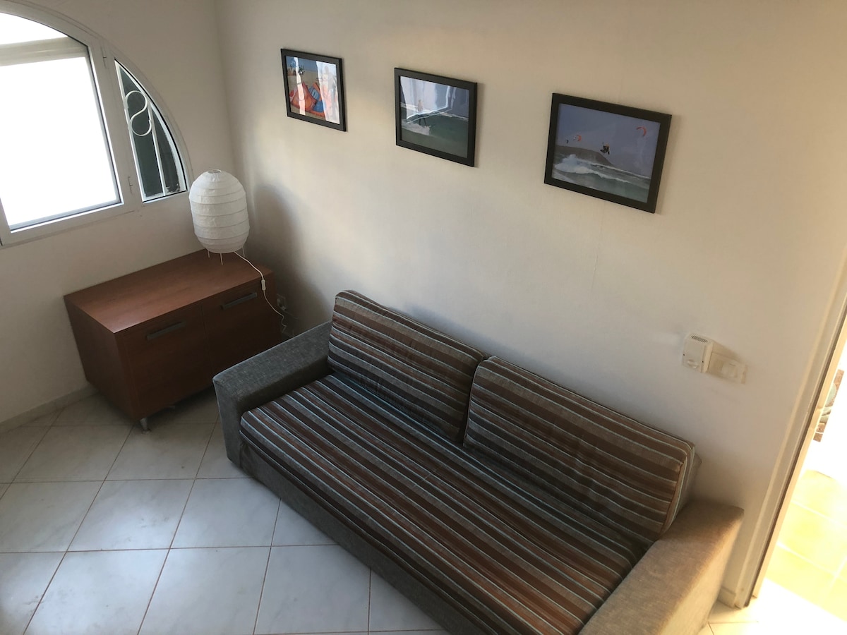 A comfortable sofa is positioned against a wall, flanked by a decorative wooden cabinet. Framed photographs of surfing activities are displayed above the sofa. Natural light enters through a window, illuminating the tiled floor.
