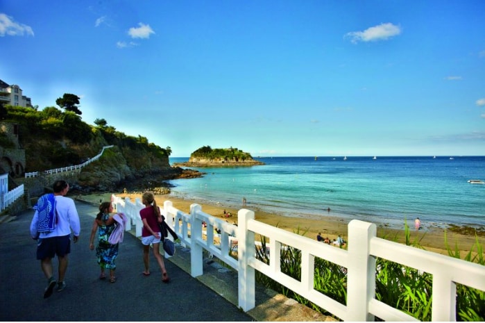 A coastal path leads toward a sandy beach, where distant figures walk along the shoreline. Gentle waves lap at the water's edge under a clear blue sky, with boats appearing in the horizon. Lush greenery and white fencing frame the scene.