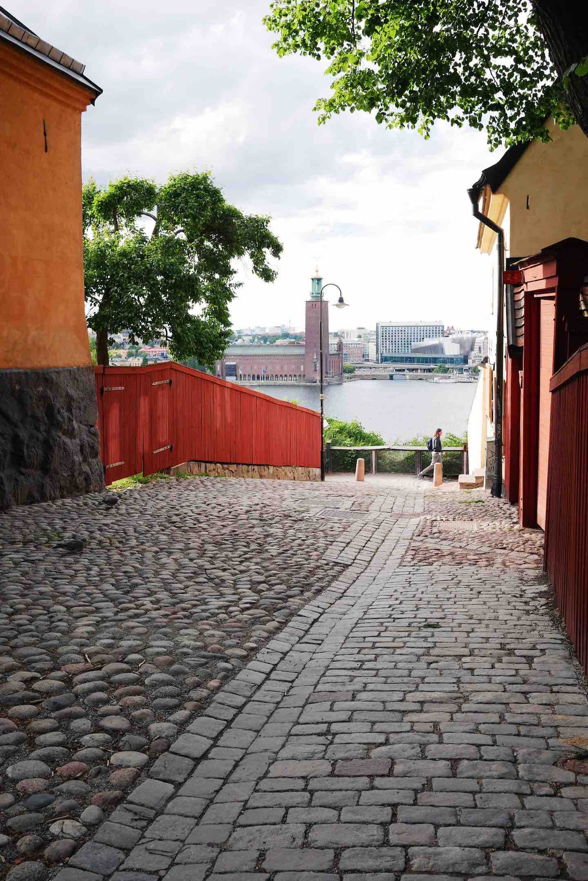 A cobblestone path descends between yellow and red buildings, leading towards a waterfront view. Trees provide shade along the sides, while a distant tower and buildings can be seen across the water, suggesting a scenic urban environment.