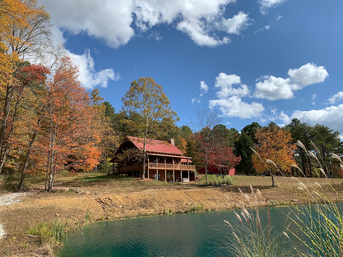 A log cabin with a red roof sits next to a tranquil pond, surrounded by trees exhibiting autumn colors. The expansive deck offers views of the scenic landscape, including colorful leaves and soft clouds in a bright sky.