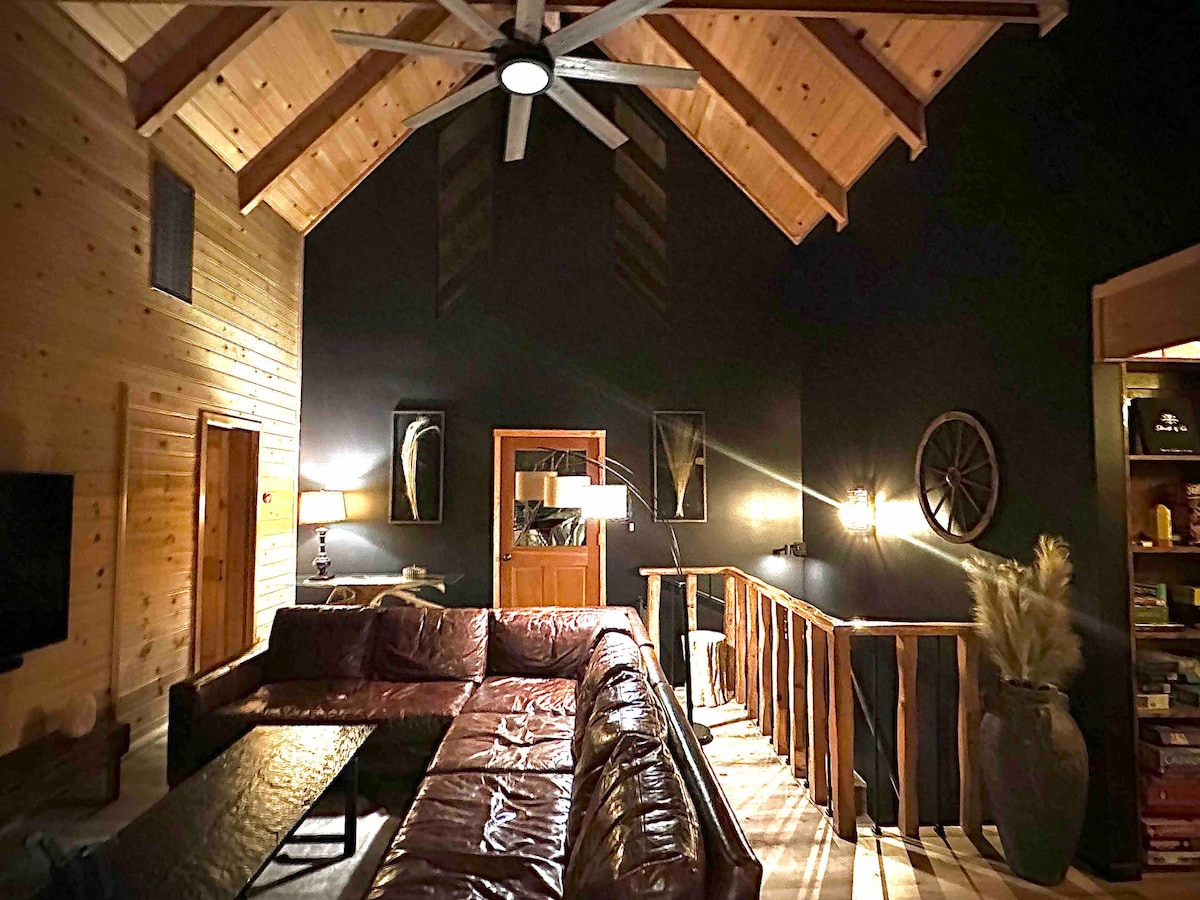 The spacious living area is highlighted by a vaulted ceiling with wooden beams and a large ceiling fan. A leather sectional sofa encircles a rectangular table, while warm lighting creates a cozy ambiance. Natural wood accents complement the dark walls, enhancing the rustic charm.