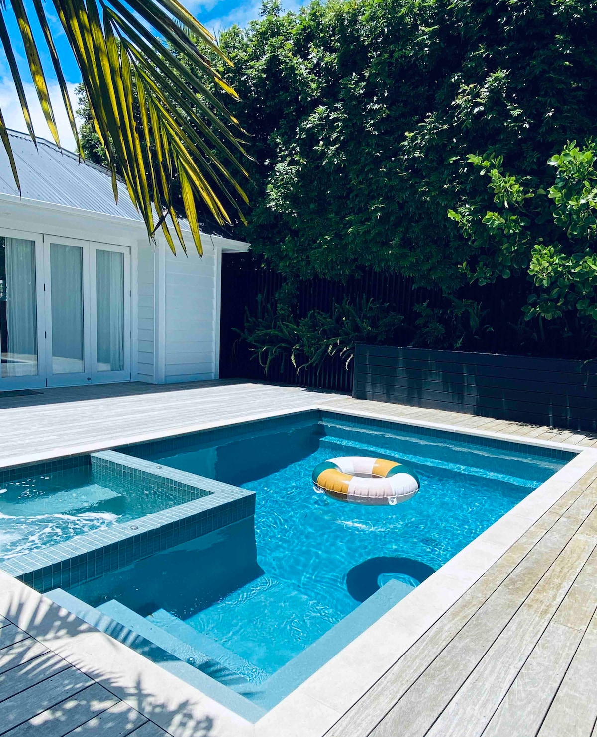 A private outdoor space features a saltwater pool surrounded by wooden decking. A spa area is integrated into the pool, while lush greenery lines the perimeter, providing a sense of tranquility. A floating life ring with bright colors is positioned within the clear blue water.
