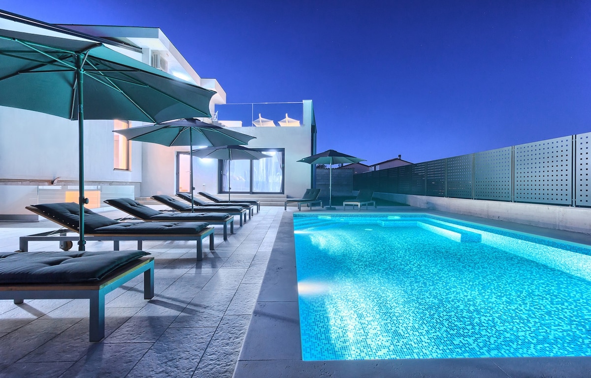 An inviting outdoor space features a swimming pool illuminated with a soft blue light, complemented by several sun loungers and large umbrellas. The surrounding area includes a modern villa, highlighted by expansive glass doors and a serene night sky.