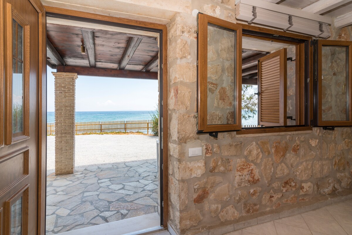 A stone entryway provides a view of the serene outdoor space and the blue waters of the Ionian Sea. The spacious area features wooden doors that open to a shaded patio, while the textured stone walls enhance the natural feel of the surroundings.