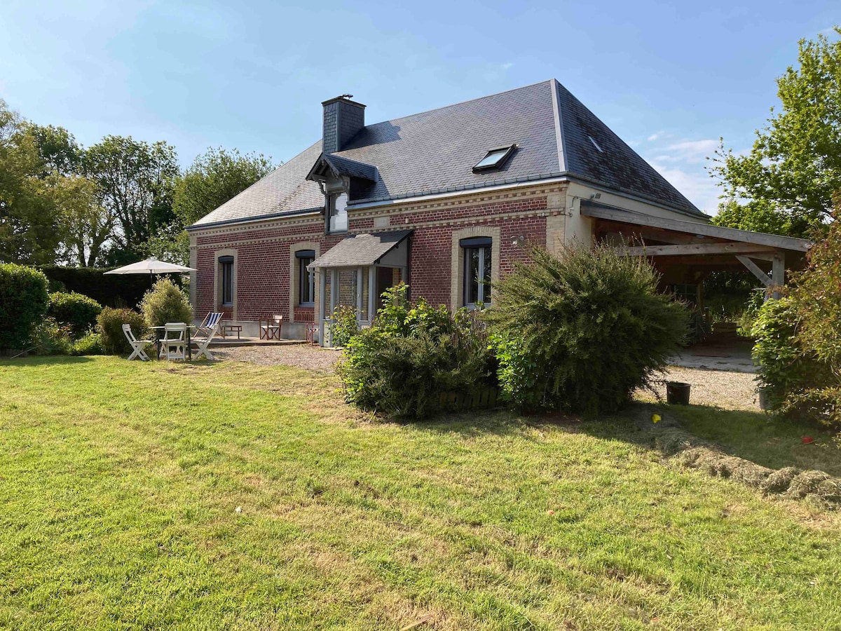 A charming house is located in a serene environment, featuring a mix of brick and stone exteriors. A spacious garden is surrounded by greenery, offering a sense of tranquility. Outdoor seating arrangements are visible, along with a shaded area for relaxation.