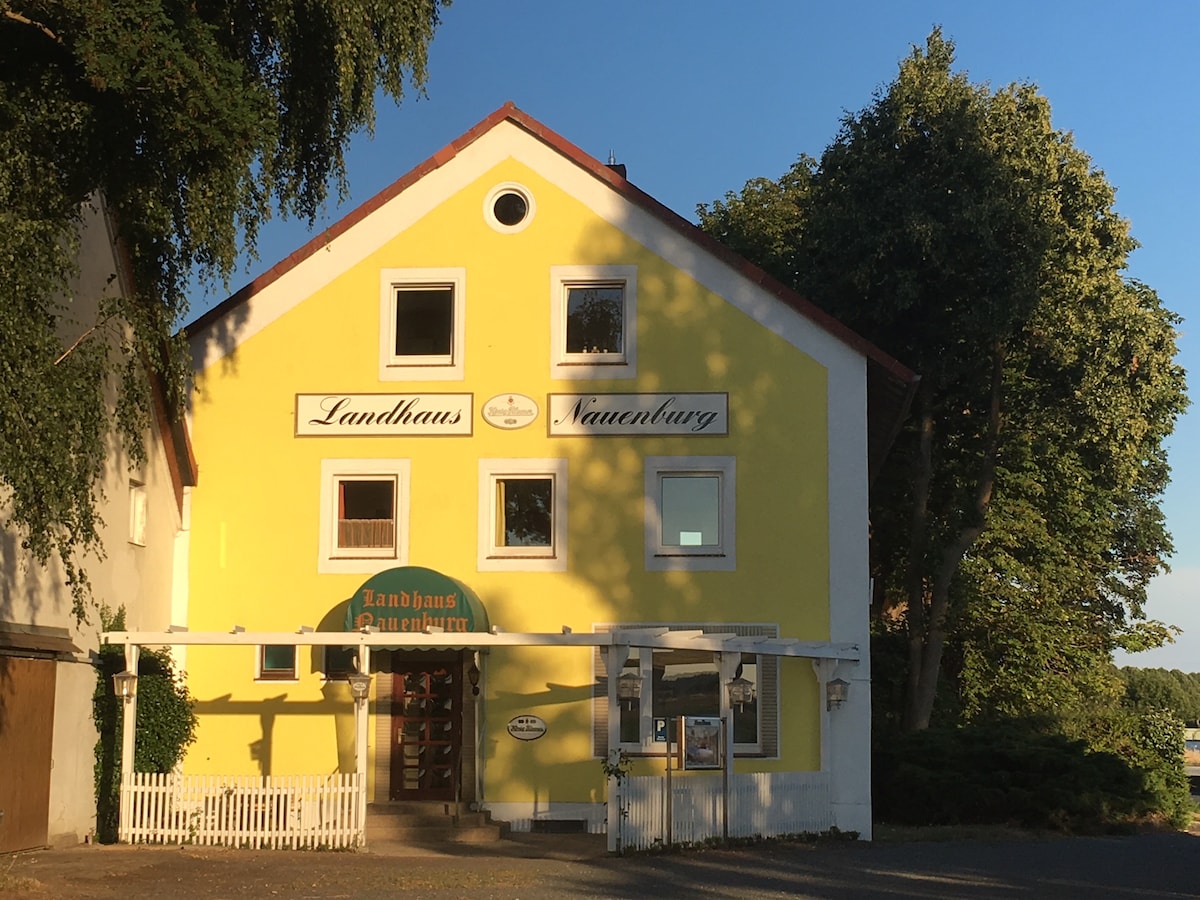 A brightly painted yellow building is showcased, featuring multiple windows with white frames and a welcoming entrance. The signage above the door indicates the property's name. Lush green trees are visible on one side, providing a natural backdrop.