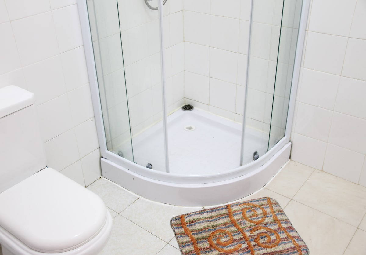 A clean bathroom features a curved glass shower enclosure and a modern toilet. A colorful mat lies on the tiled floor in front of the shower, adding a touch of warmth to the space. White tiles cover the walls, enhancing the bright and airy feel.