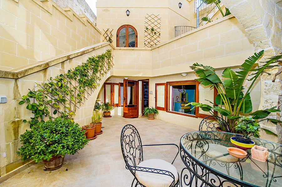 A private courtyard is captured, featuring a round glass table and black iron chairs. Lush greenery surrounds the area, with potted plants positioned along the walls. The entrance to the suite is visible through large wooden doors, illuminated by natural light filtering into the space.