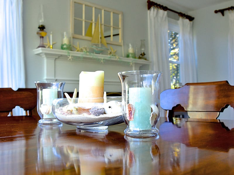 A wooden dining table is set with a centerpiece featuring three candles surrounded by seashells. Two glass holders with candles are placed on either side. A bright, airy atmosphere is created by natural light streaming through large windows with white curtains.