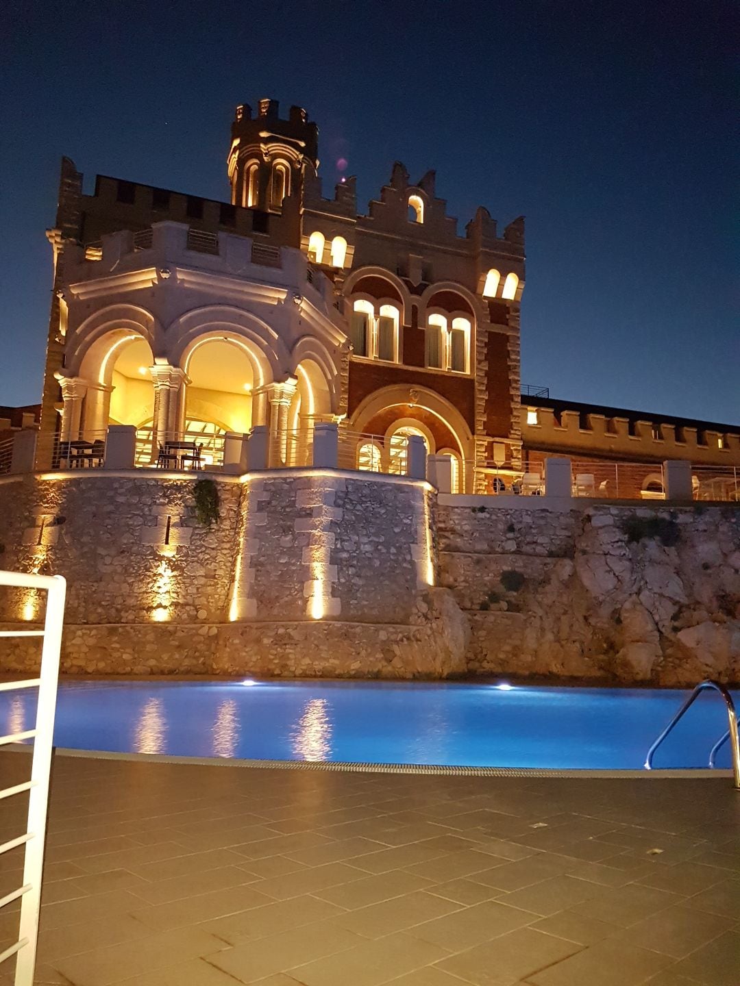 The image captures a beautifully illuminated villa at night, showcasing intricate architectural details. Soft lighting highlights the stone facade and elegant arches. A serene pool reflects the glowing structure, creating a peaceful outdoor environment.