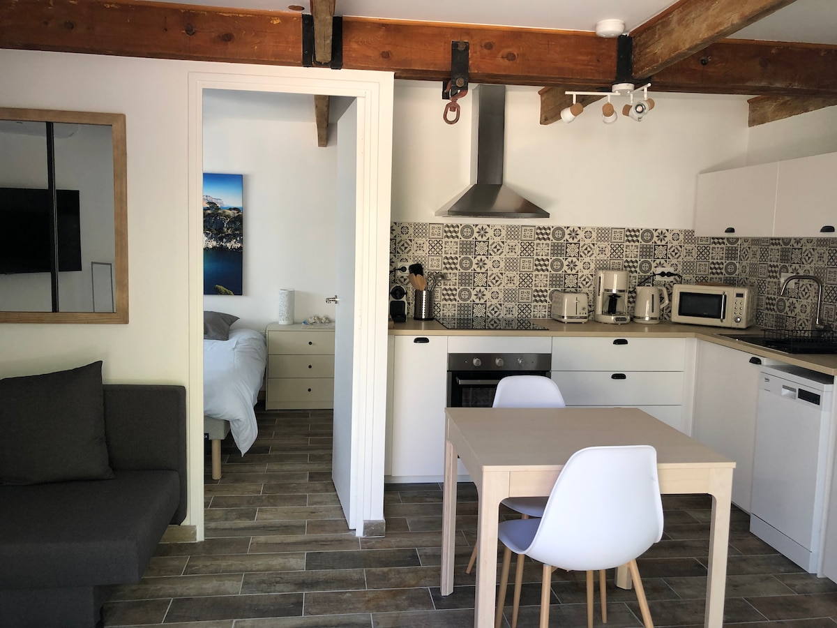 The image features a modern kitchen area with patterned tiles and a sleek design. A dining table with white chairs is situated at the center, while a grey sofa is positioned nearby. A doorway leads to a well-lit sleeping area, partially visible in the background.
