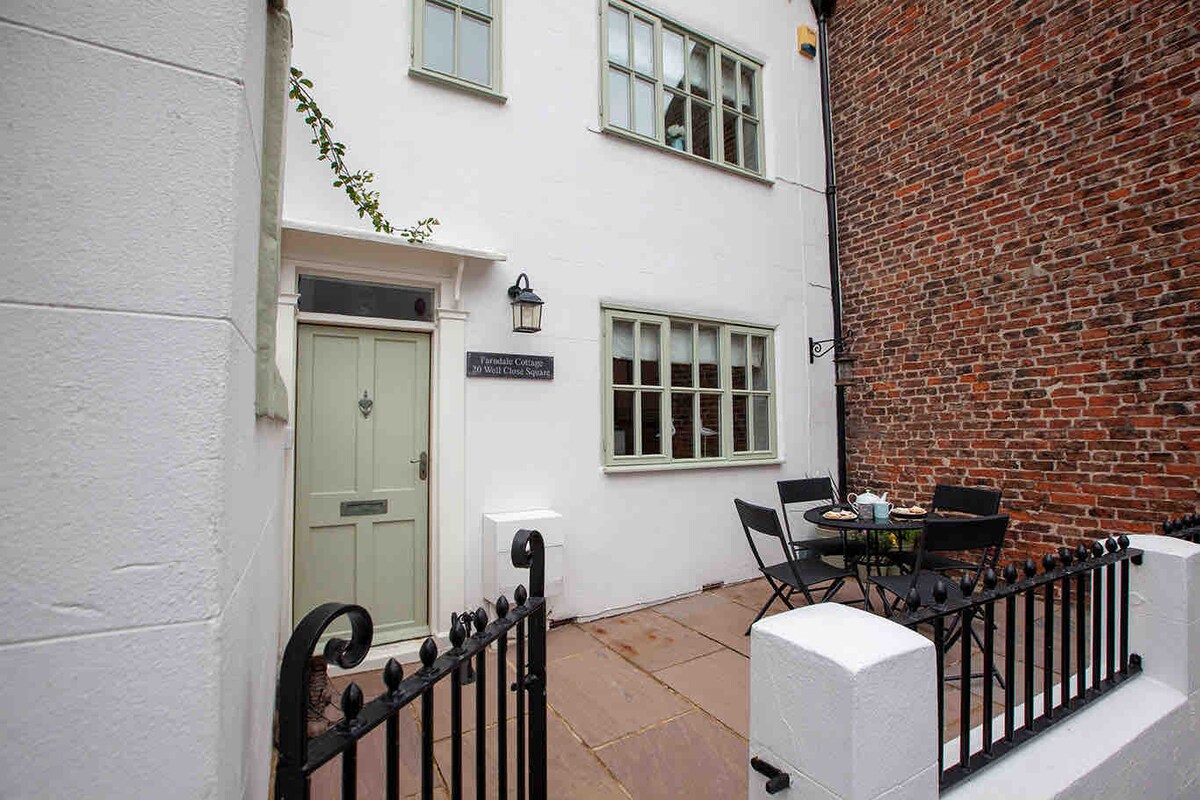 The exterior of the cottage is presented with crisp white walls and a welcoming green door. A small outdoor seating area is arranged with a round table and chairs, complemented by a stone pathway and surrounding brick wall.