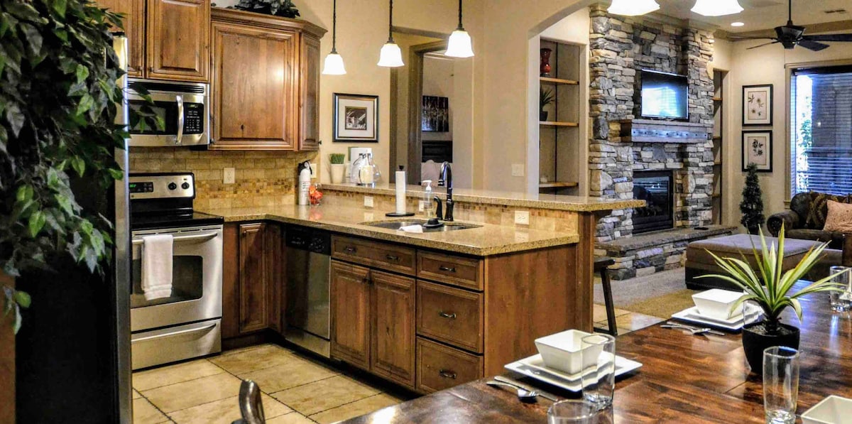 A modern kitchen features wooden cabinetry and stainless-steel appliances. A spacious granite countertop provides ample workspace. Under-cabinet lighting highlights the area, while a dining table set for four sits nearby. A cozy living area is visible in the background, equipped with a stone fireplace and television.
