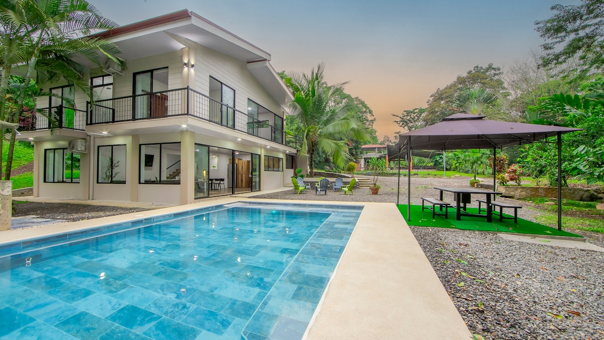The image showcases a modern two-story house with multiple balconies, set against a backdrop of lush tropical vegetation. A private swimming pool reflects the surroundings, while a gazebo and outdoor dining area can be seen nearby, offering spaces for relaxation and enjoyment.