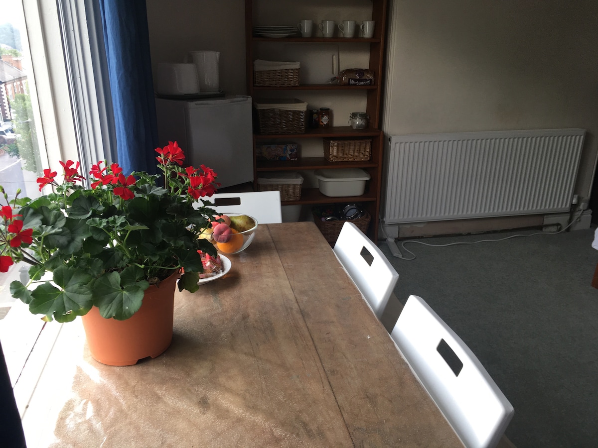 A dining area is shown with a wooden table and four white chairs. A potted plant with red flowers sits on the table beside a bowl of fruit. In the background, a shelving unit displays various baskets and dishware, while a radiator is positioned nearby.