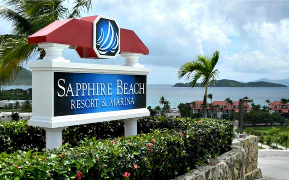 The image features a well-maintained sign for Sapphire Beach Resort & Marina, prominently displayed against a backdrop of lush greenery and vibrant tropical plants. The serene blue waters and hills are visible in the distance, providing a glimpse of the coastal beauty surrounding the resort.