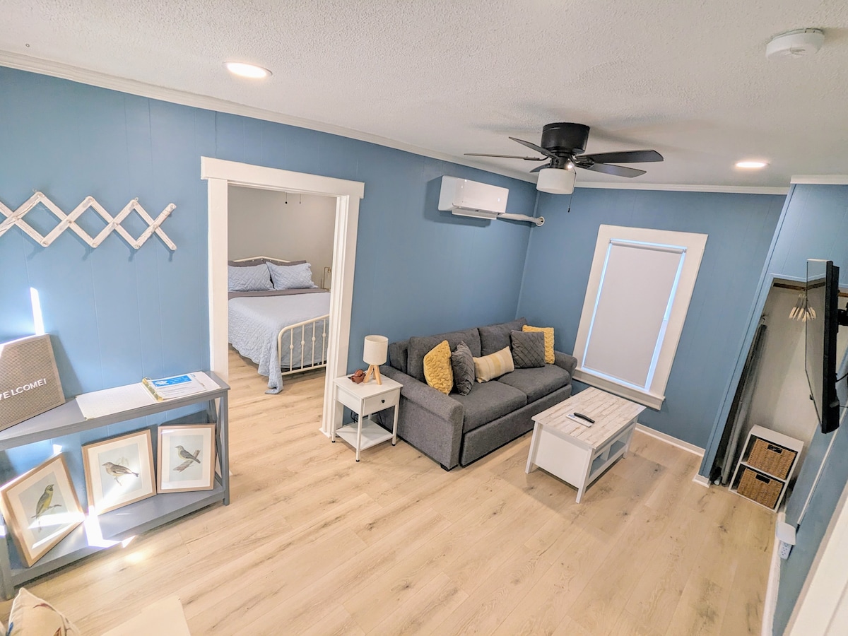 A serene living area is presented with light blue walls and ample natural light. A gray sofa with yellow-accented pillows faces a white coffee table. A white decorative wall feature adds charm, with a doorway leading into a bedroom visible in the background.