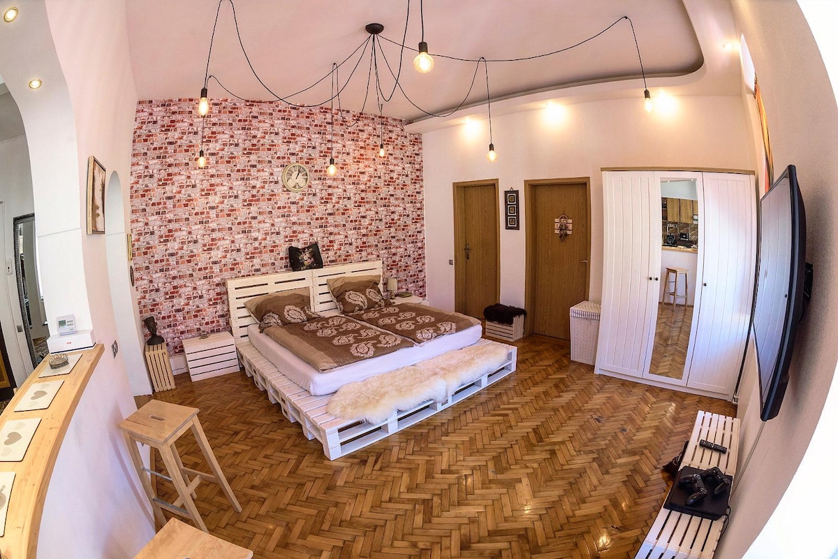 A spacious bedroom is highlighted by a cozy double bed adorned with neutral-toned bedding. A feature wall of exposed brick adds texture, while a vintage clock complements the decor. A large mirror and seating area are visible, with stylish pendant lights providing illumination.