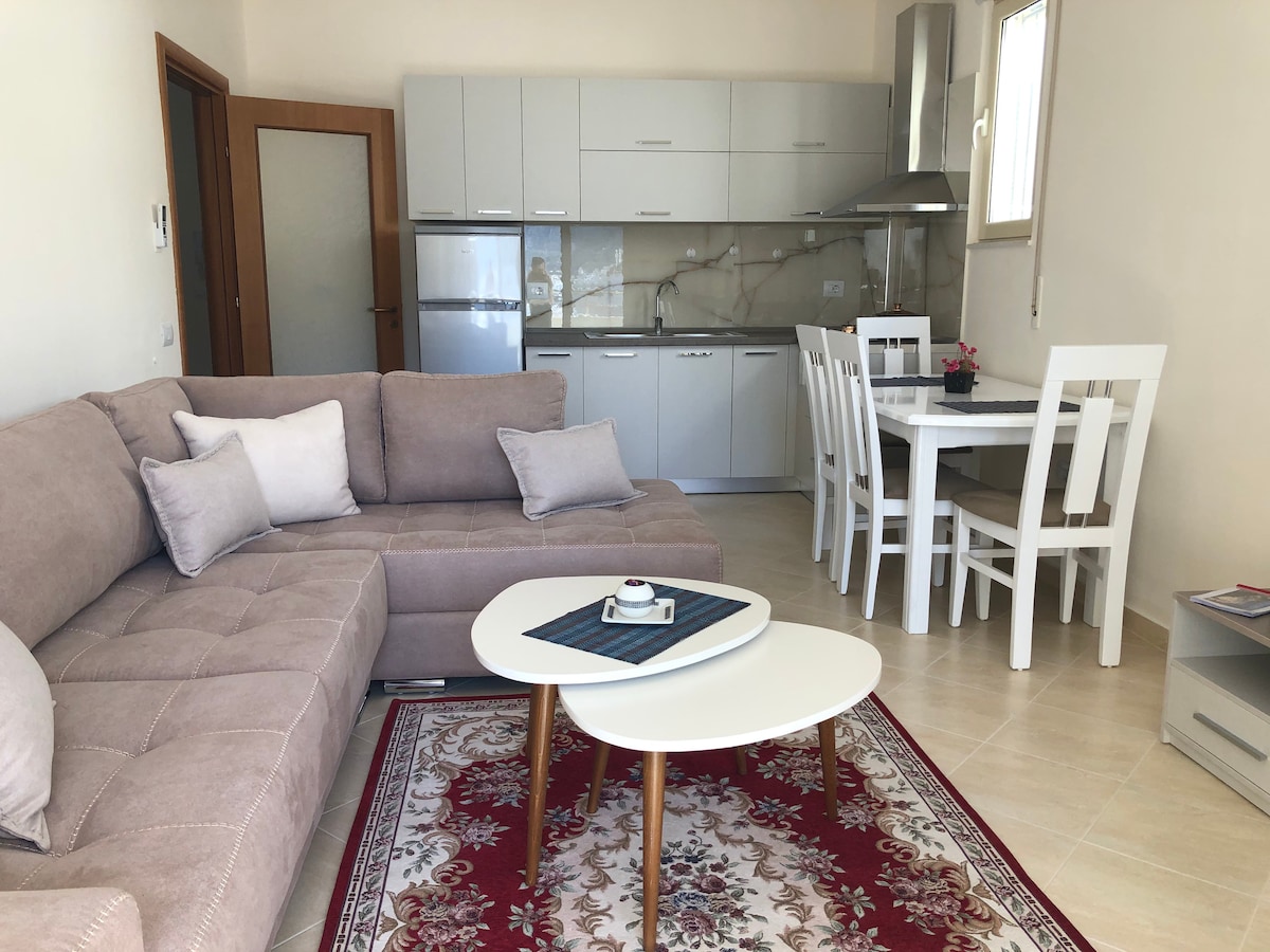 A modern living area is presented, featuring a light-colored sectional sofa and a small coffee table. A dining table with white chairs is visible in the background, alongside a kitchen area with sleek cabinets and appliances. Natural light enters through a window.